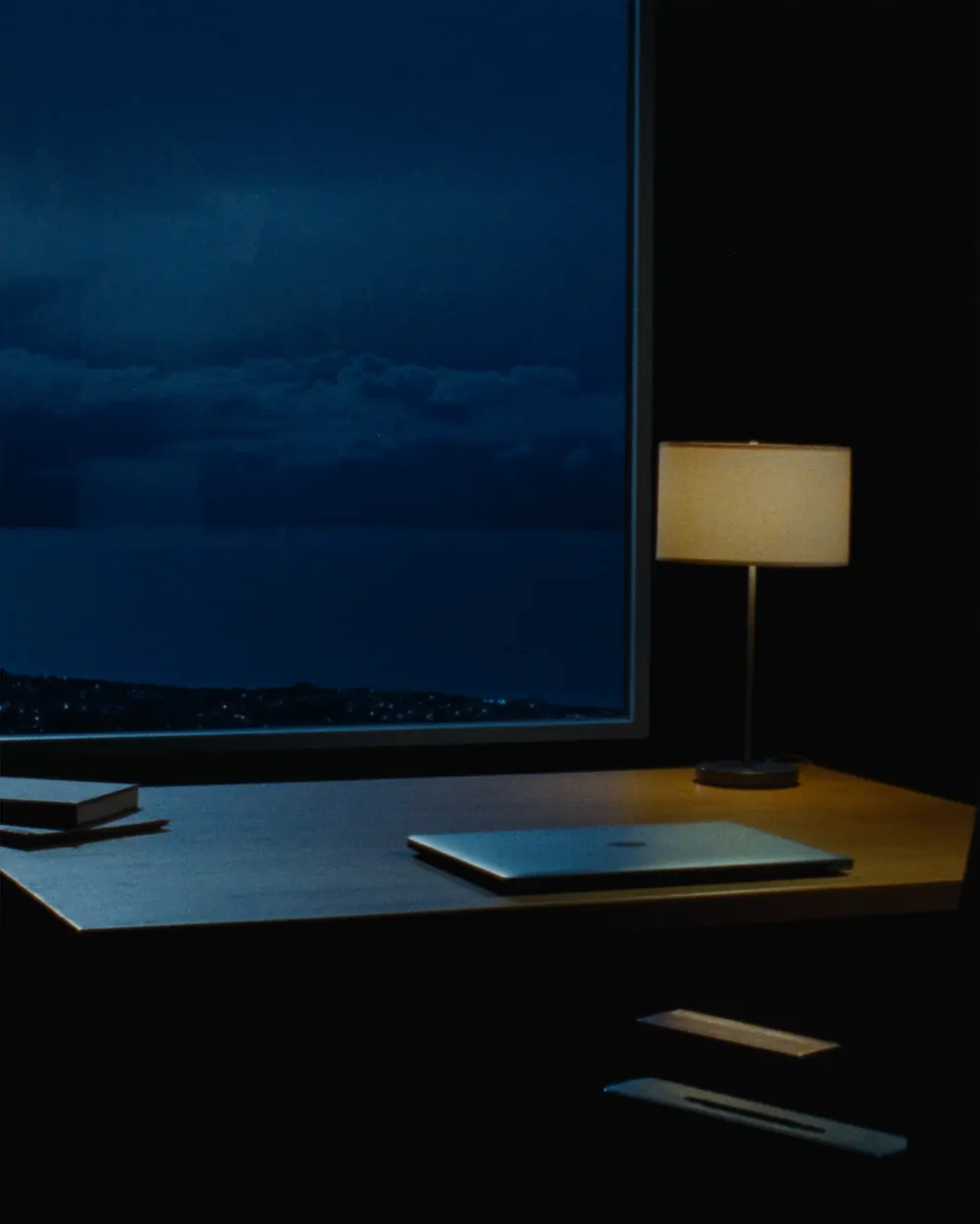 A desk at night with a warm lamp and a window onto a moonlit sea of clouds